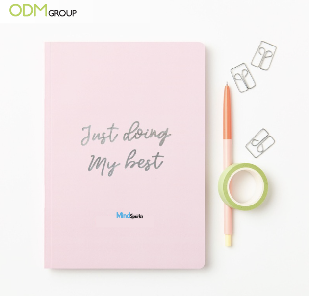 Pink notebook with the phrase "Just doing My best" on the cover, alongside a pen, tape, and paper clips.
